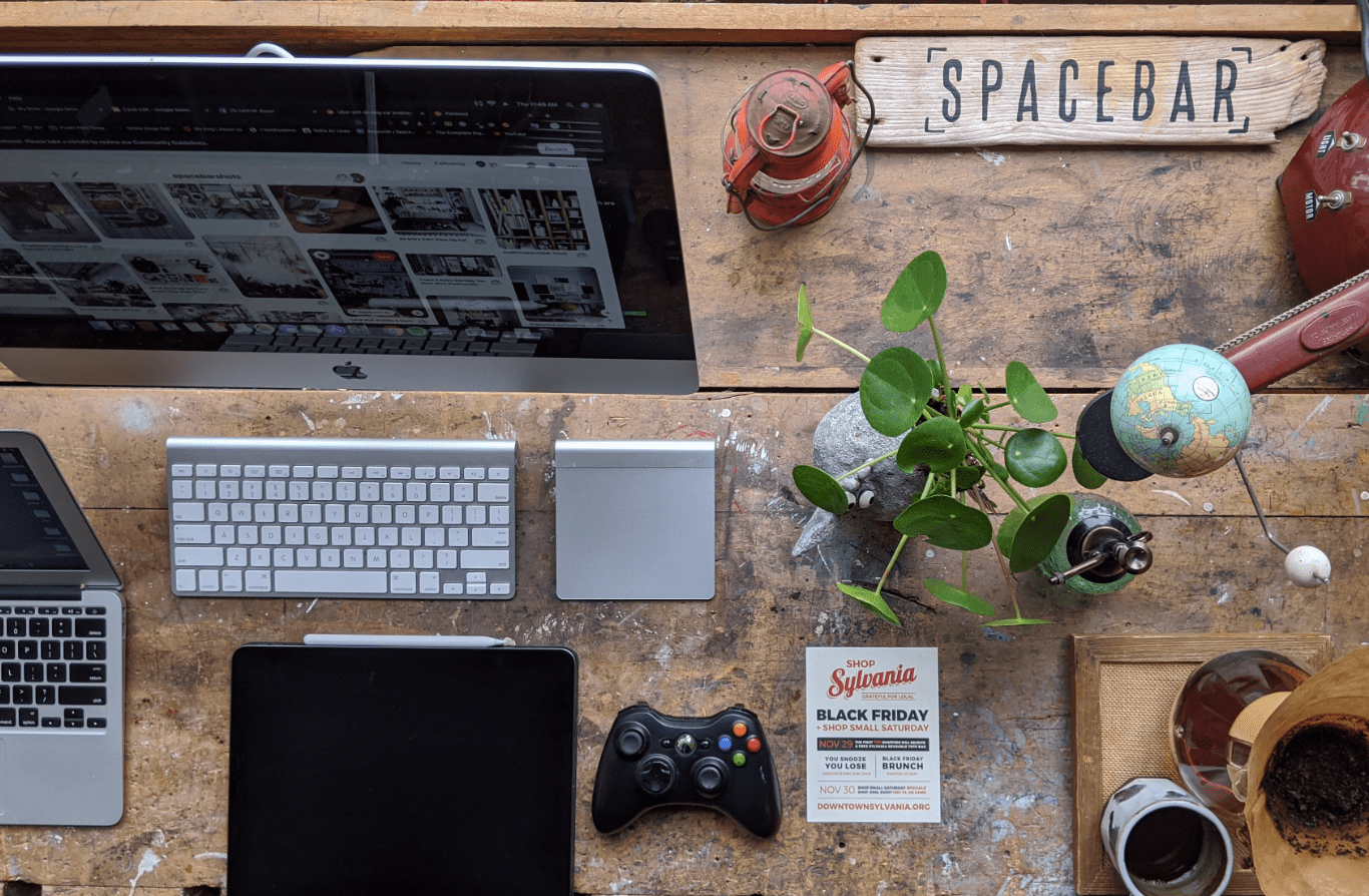 The original creative workspace: a wooden desk with computers, a camera, a globe, and a Shop Sylvania flyer.